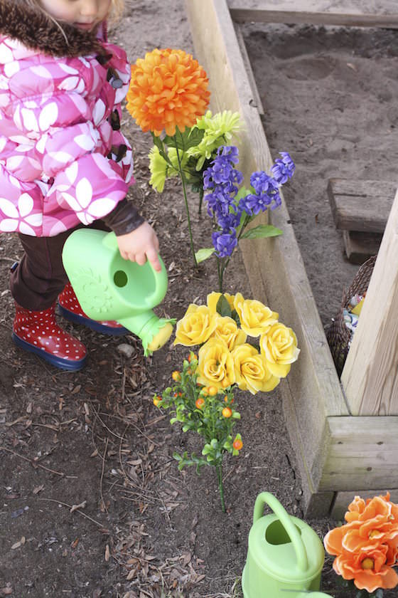 preschooler watering artificial flowers beside sandbox