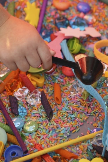 Preschooler scooping coloured rice in a sensory bin filled with assorted colourful items