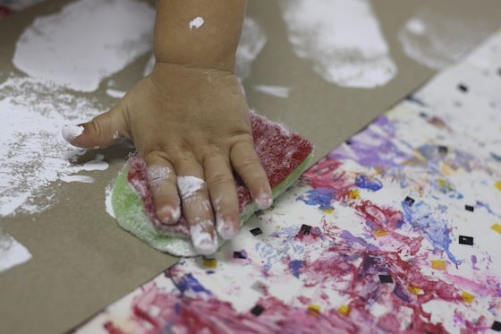 toddler hand pressing painted sponge on cardboard