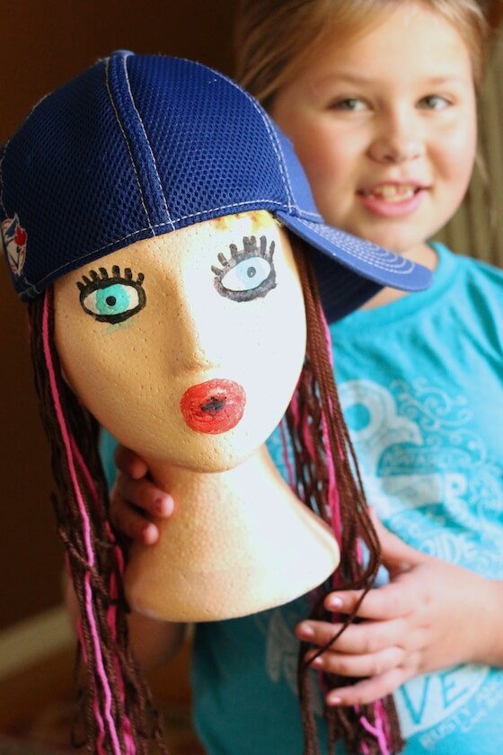 girl holding styrofoam head decorated and wearing ball cap