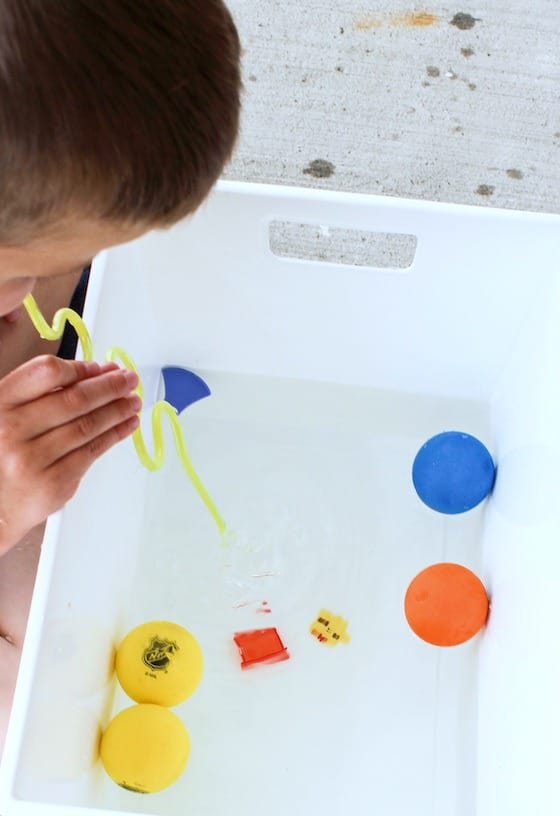 child blowing toys across water with drinking straw